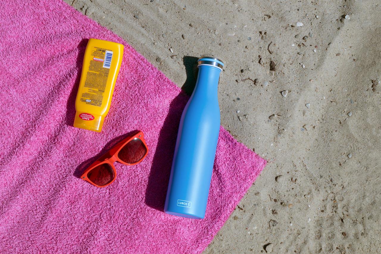 Auf dem Bild liegt eine azurblaue Isolierflasche aus Edelstahl (0,75l) auf einem pinken Handtuch am Strand. Daneben befinden sich eine gelbe Sonnencreme und eine rote Sonnenbrille.