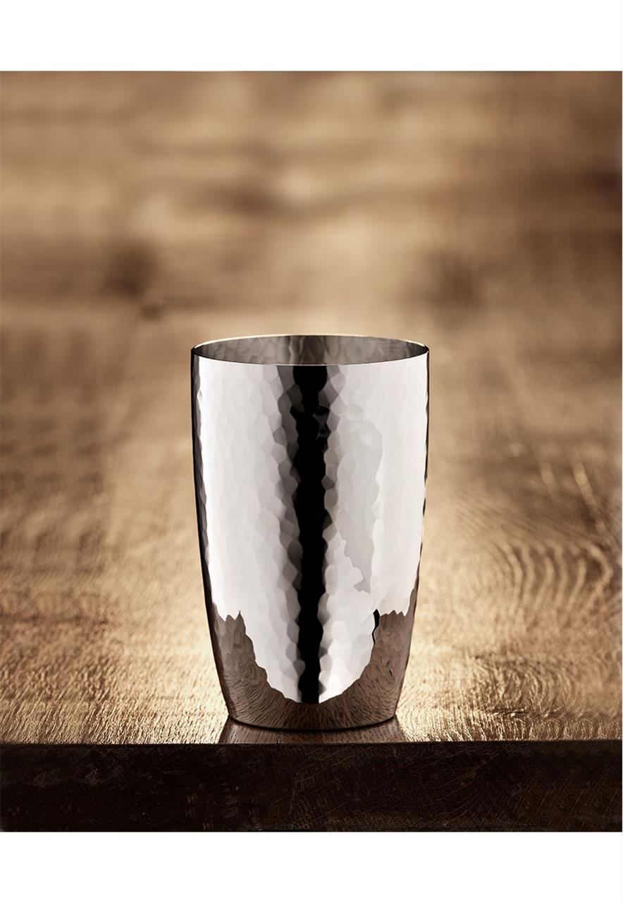Das Bild zeigt den Martelé 90 Longdrinkbecher aus glänzendem Metall mit einer gehämmerten Oberfläche. Der Becher steht auf einer Holzfläche und reflektiert das Licht elegant.