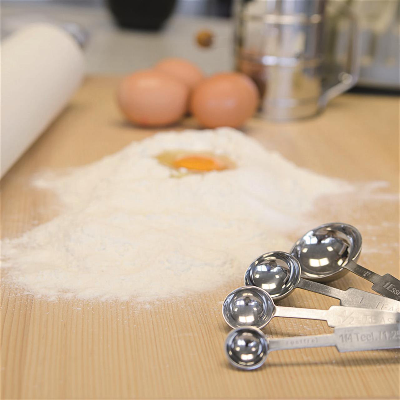 Im Vordergrund des Bildes liegt ein Messlöffel-Set aus Metall auf einer Holzoberfläche. Im Hintergrund sind Mehl, ein aufgeschlagenes Ei und einige Eier zu sehen, was auf eine Backvorbereitung hinweist.