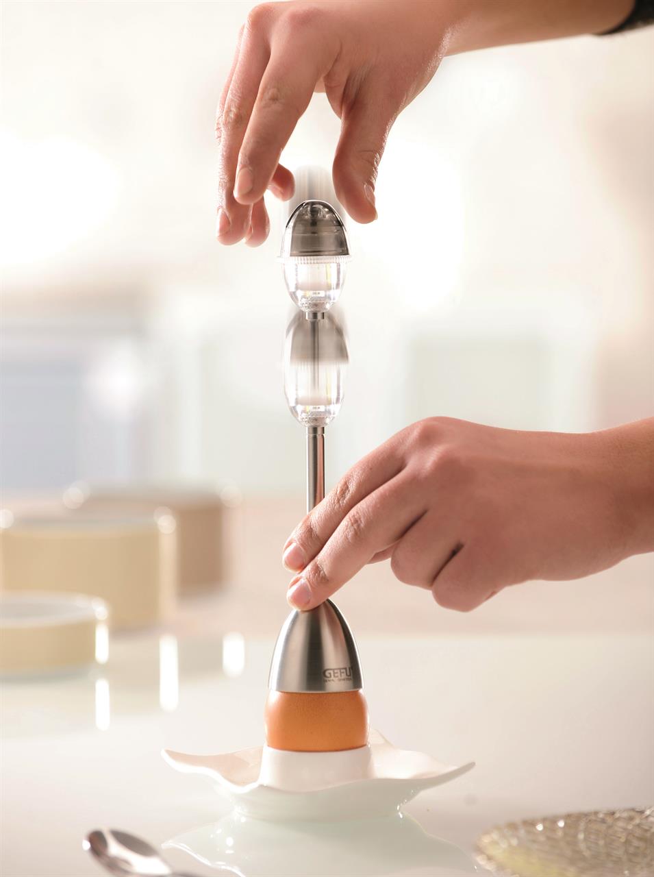 Das Bild zeigt eine Person, die einen Eierköpfer benutzt, um ein Ei zu öffnen. Das Gerät steht auf dem Ei und wird mit einer Hand bedient.