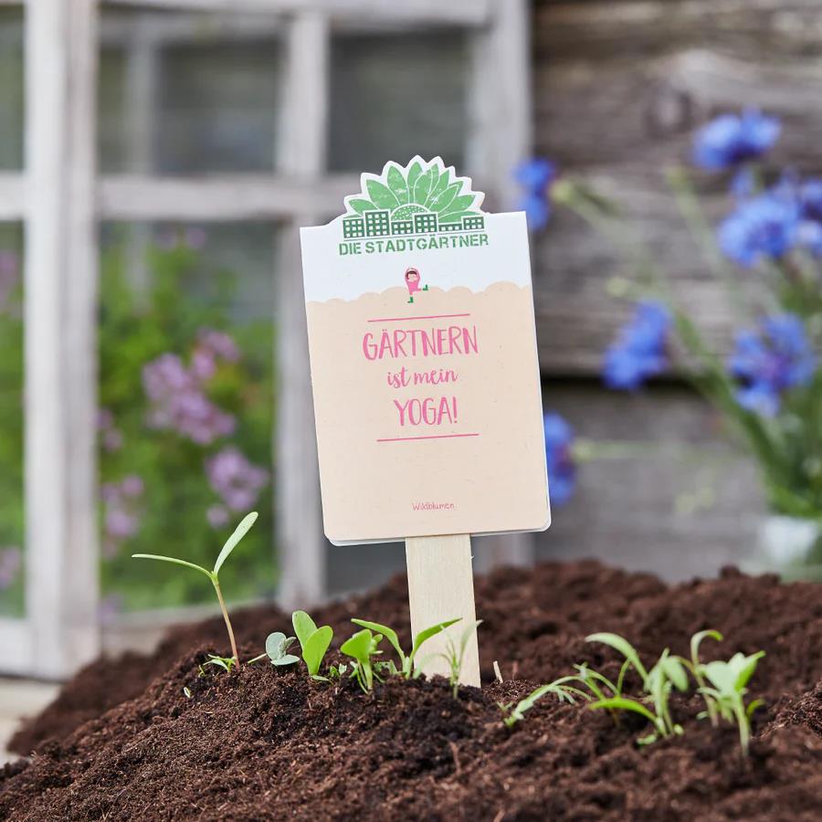 Das Bild zeigt ein Samentütchen mit der Aufschrift "Gärtnern ist mein Yoga!" im Beet. Im Vordergrund sieht man junge Pflänzchen, im Hintergrund blühen Blumen.
