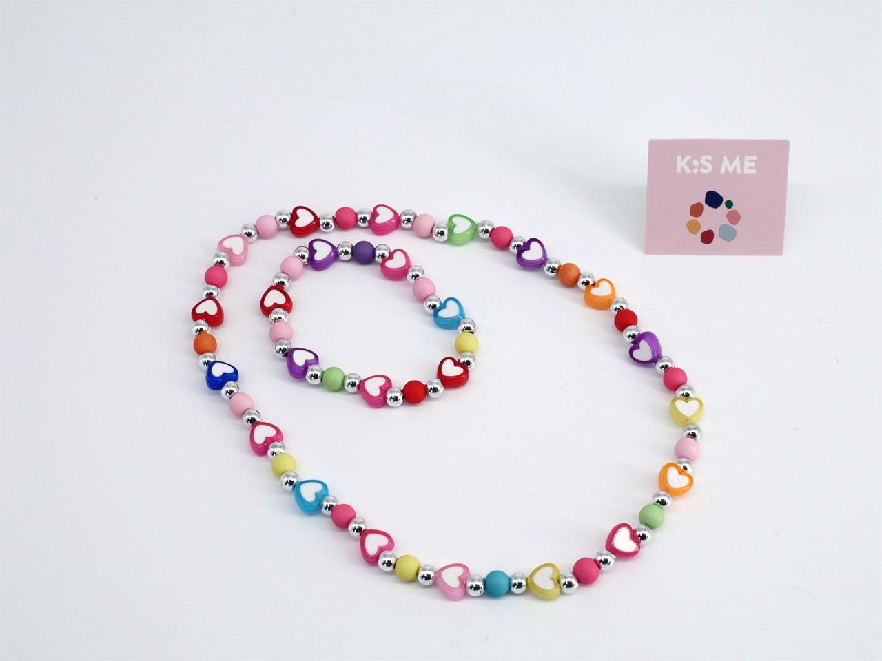 Das Bild zeigt ein Set aus einer Kette und einem Armband mit bunten Herzperlen. Im Hintergrund steht eine kleine Karte mit der Aufschrift "K:S ME".