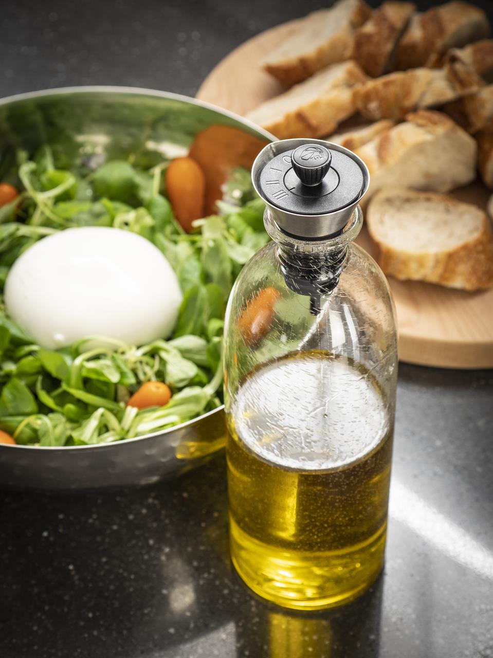 Im Vordergrund ist der Öl- und Essigausgießer Ciro auf einer Flasche zu sehen. Im Hintergrund stehen ein frischer Salat und aufgeschnittenes Brot bereit.