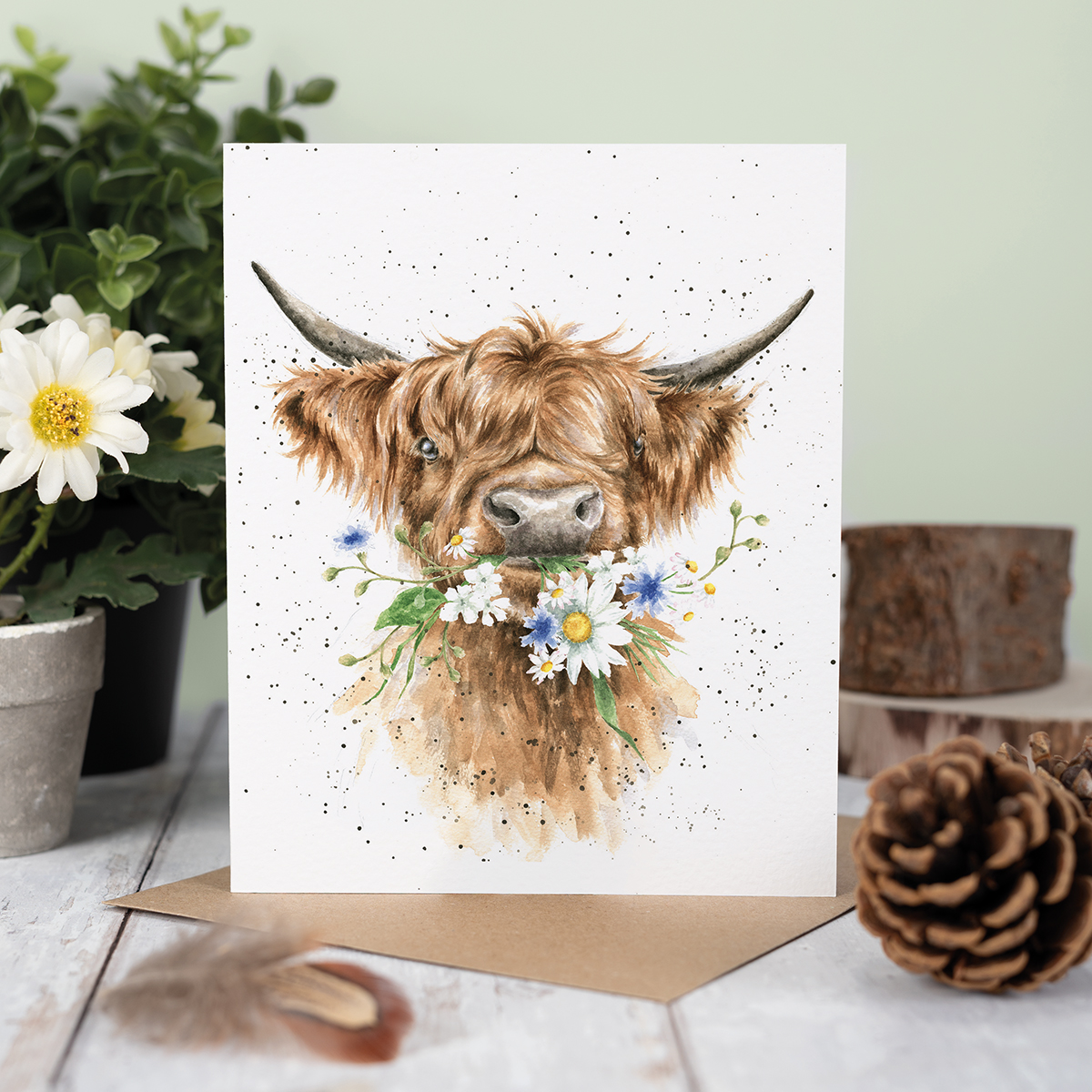 Die Karte zeigt eine niedliche Hochlandkuh mit Blumen im Maul. Im Hintergrund sind Pflanzen und Naturdekoration zu sehen.