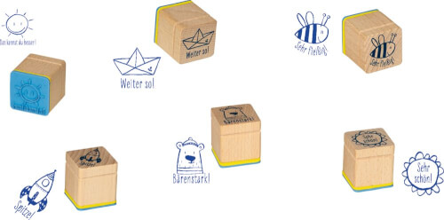 Auf dem Bild sind mehrere Holzstempel mit unterschiedlichen Motiven und deutschen Sprüchen zu sehen. Jeder Stempel hinterlässt ein einzigartiges Bild, wie ein Papierboot, eine Biene oder eine Sonne, zusammen mit kurzen Lobworten.