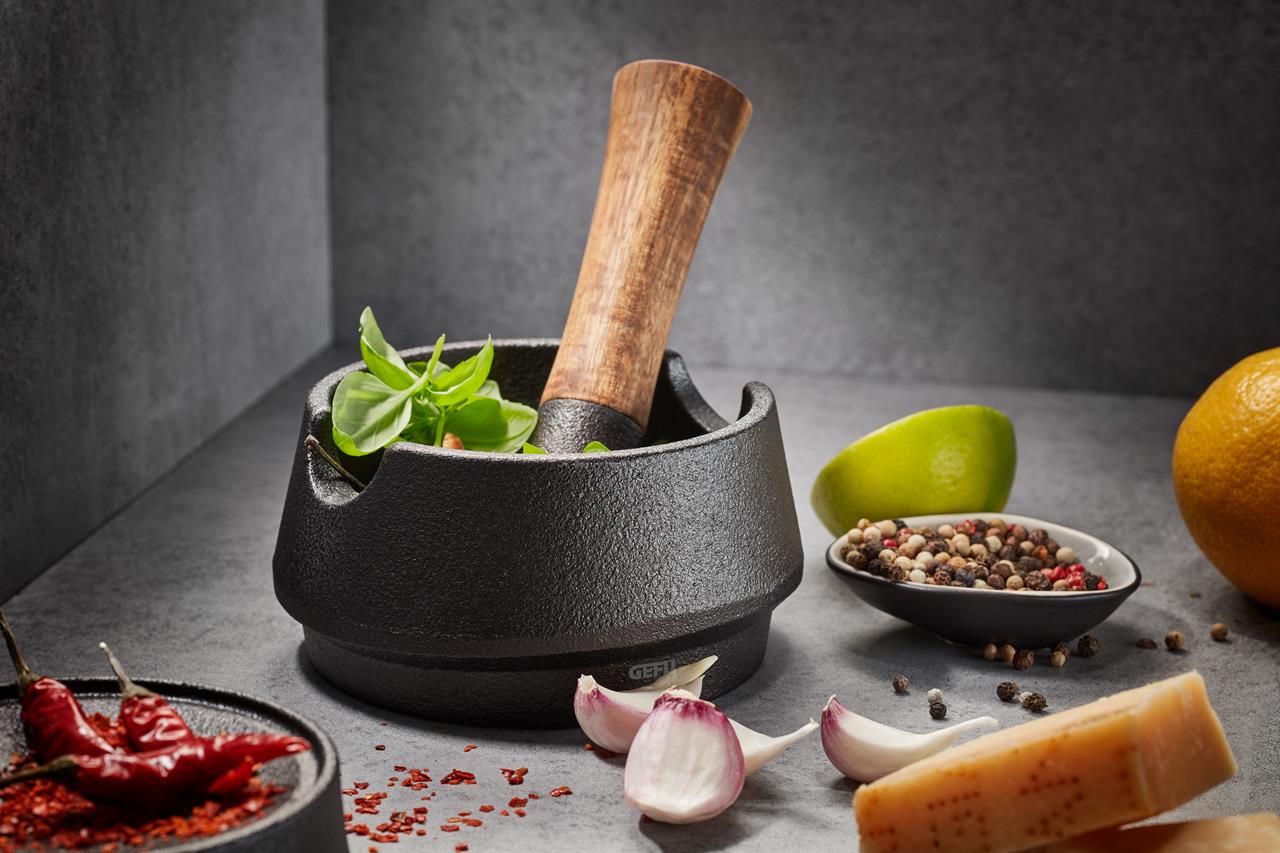 Der Gussmörser X-PLOSION® steht im Mittelpunkt des Bildes, gefüllt mit frischen Kräutern und einem Holzstößel. Drumherum liegen Gewürze, Knoblauch, Käse und Zitrusfrüchte, die eine gemütliche Kochatmosphäre schaffen.