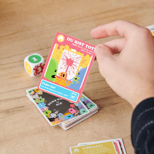 Eine Hand hält eine bunte Spielkarte mit der Aufschrift "DU BIST TOT!" über einem Stapel anderer Karten. Im Hintergrund liegt ein Würfel mit Symbolen auf einem Holztisch.