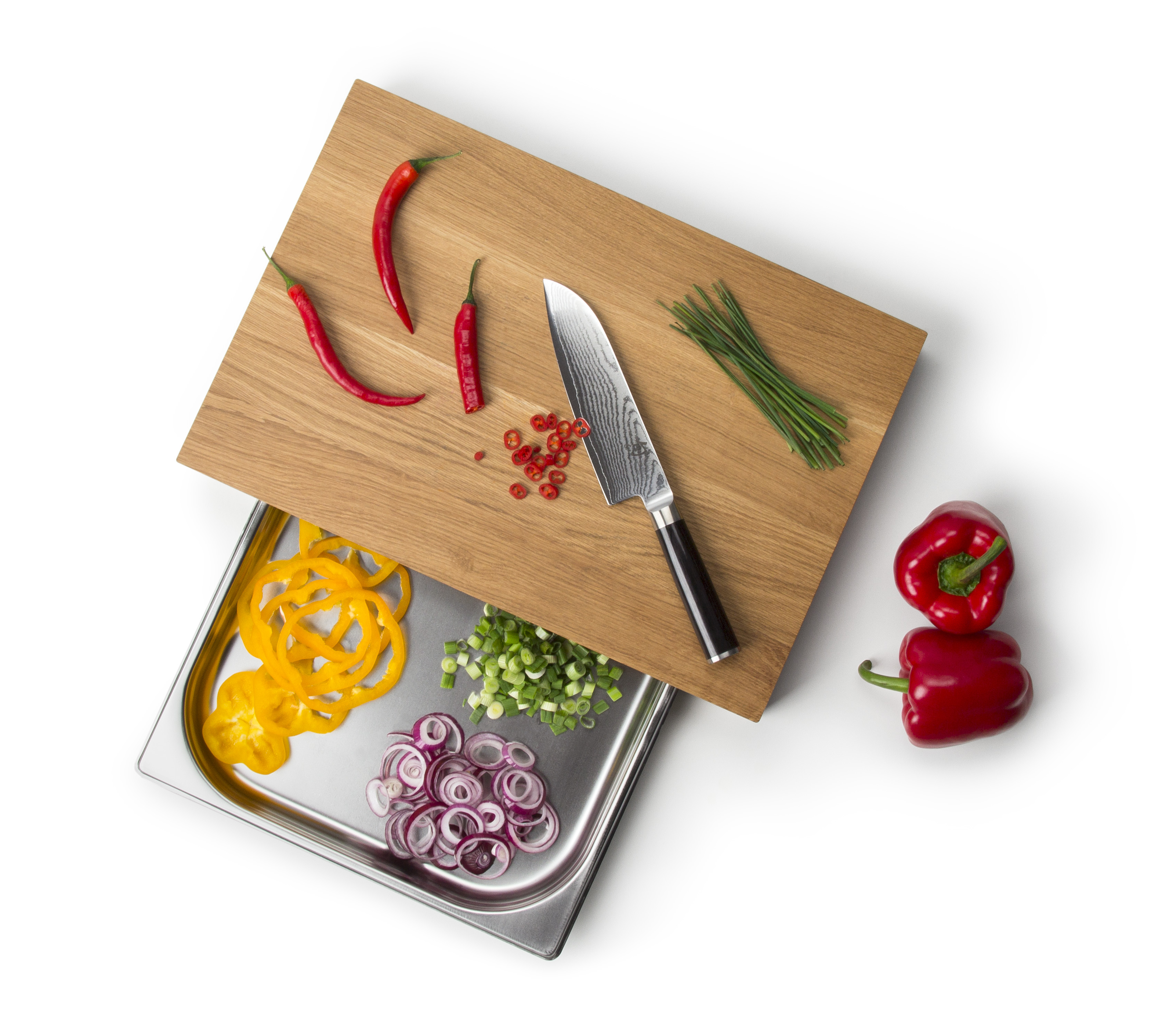 Auf dem Bild sieht man ein Schneidebrett mit Chilischoten, Schnittlauch und einem Messer. Darunter befindet sich eine Metallschale mit geschnittenen Paprika, Frühlingszwiebeln und roten Zwiebeln, daneben liegen zwei rote Paprika.
