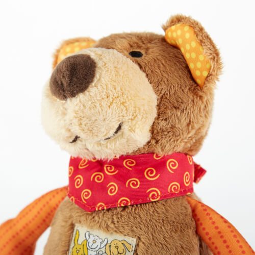 Auf dem Bild ist ein brauner Plüschbär mit orangefarbenen Ohren und einem roten Halstuch mit gelbem Muster zu sehen. Der Bär hat ein freundliches Gesicht und trägt einen kleinen Aufnäher mit Tiermotiven auf dem Bauch.