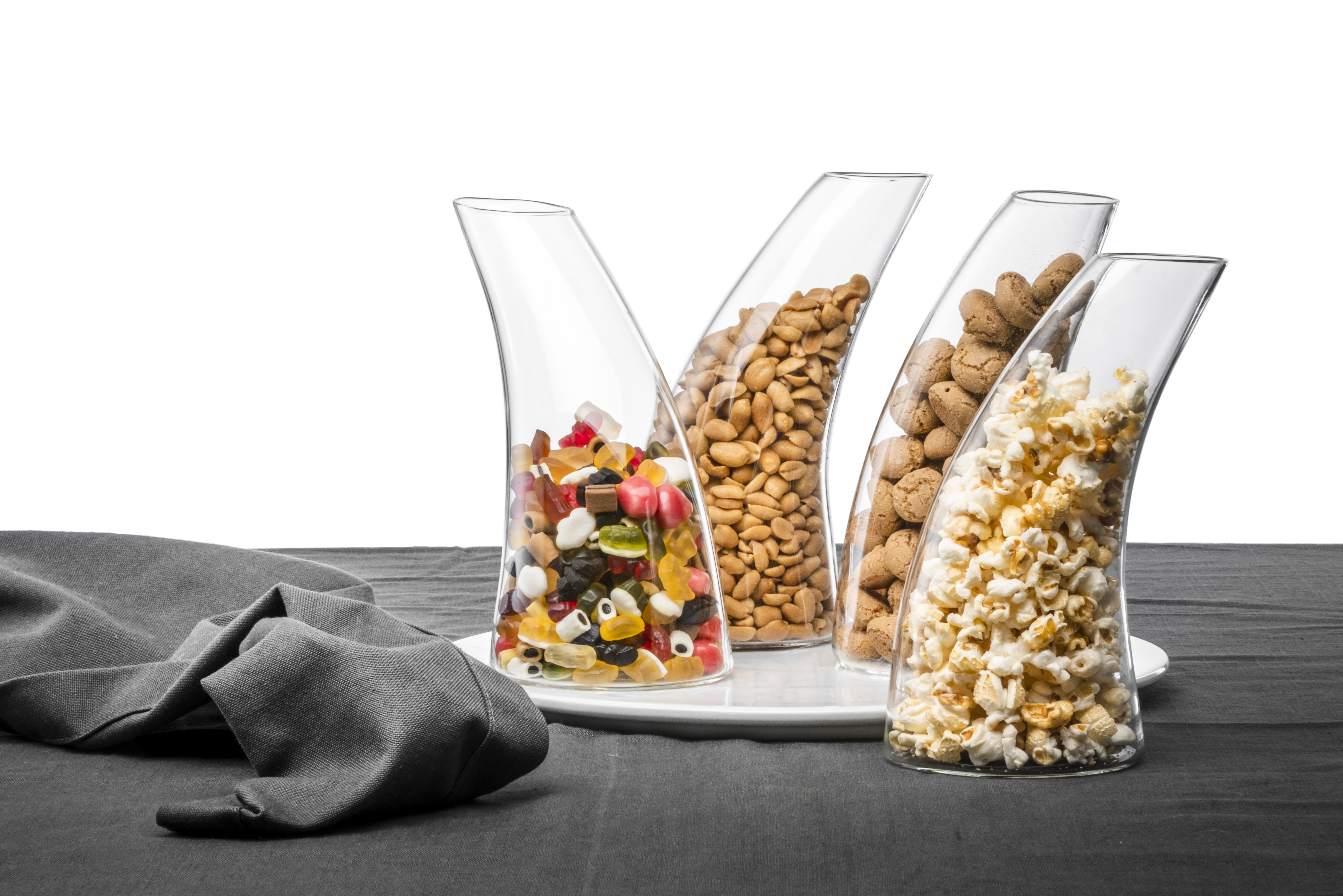 Vier schräge Glasbehälter sind nebeneinander auf einem weißen Tablett aufgestellt. Sie sind mit Gummibärchen, Erdnüssen, Keksen und Popcorn gefüllt.