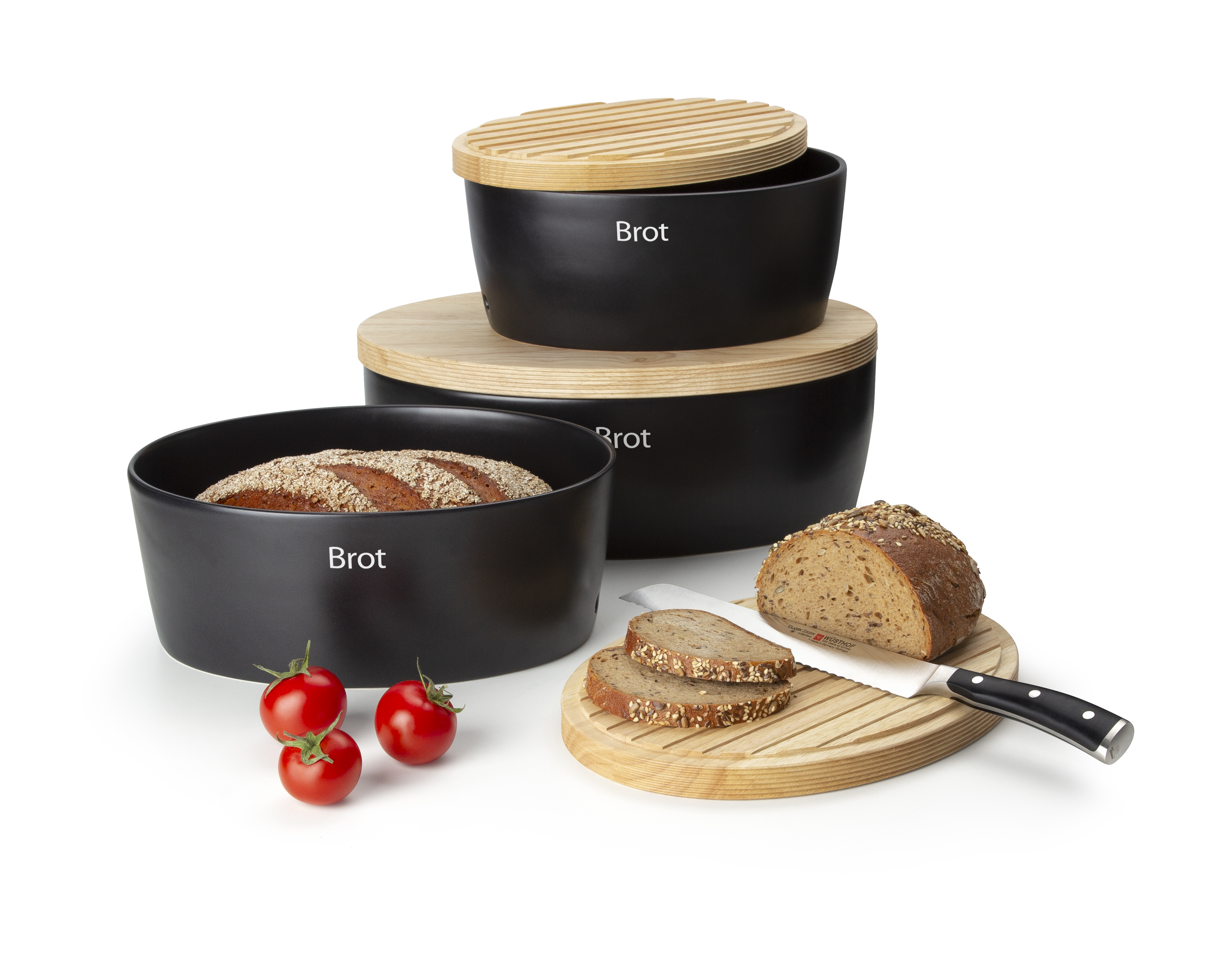 Auf dem Bild sind drei schwarze Brotdosen mit Holzdeckeln zu sehen, die das Wort "Brot" darauf haben. Daneben liegen einige geschnittene Brotscheiben, ein Messer, drei Tomaten und ein runder Holzuntersetzer.