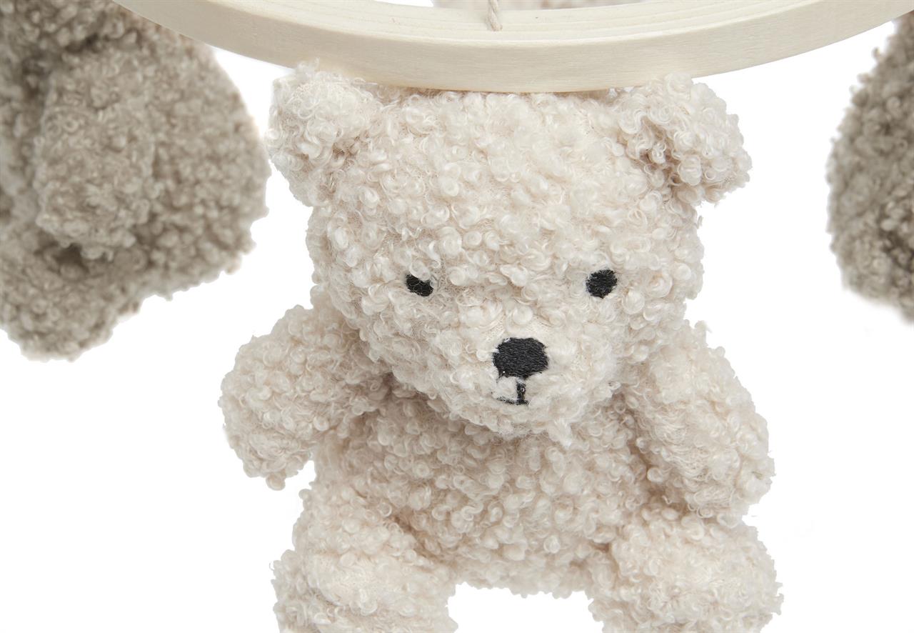 Das Bild zeigt einen weichen, cremefarbenen Teddybär, der an einem Baby-Mobile hängt. Der Teddybär besteht aus flauschigem Stoff und hat gestickte Augen und eine Nase.