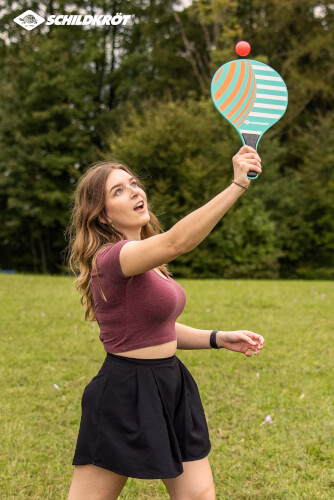 Eine junge Frau spielt draußen mit einem bunten Schläger und einem kleinen Ball. Sie steht auf einer Wiese vor einer Baumkulisse.