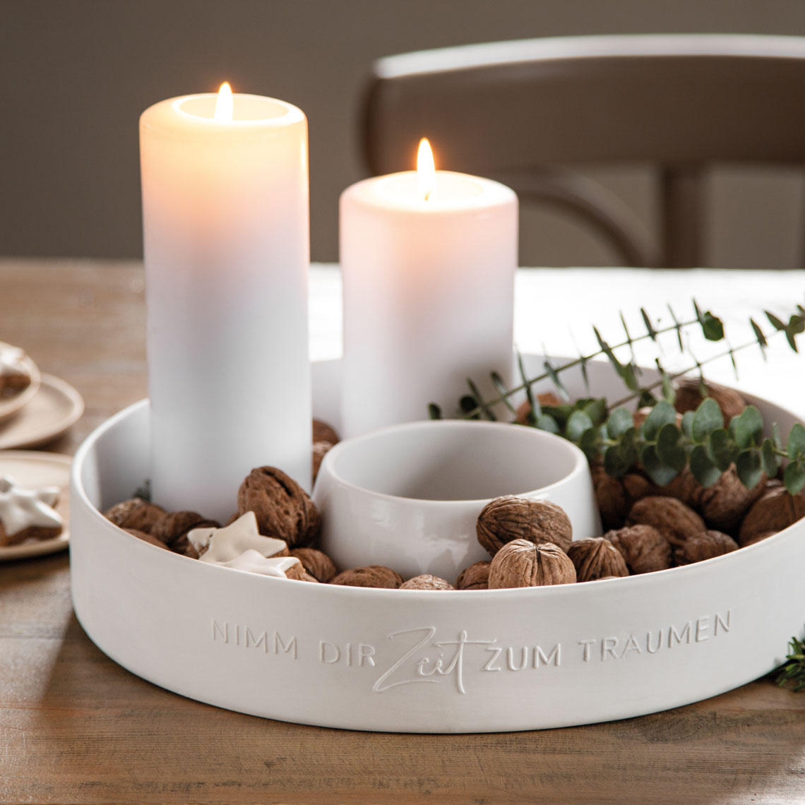 Auf dem Bild sind zwei brennende weiße Kerzen in einem runden weißen Teller zu sehen, der mit Walnüssen und grünen Zweigen dekoriert ist. Auf dem Rand des Tellers steht der Satz: "NIMM DIR ZEIT ZUM TRÄUMEN."