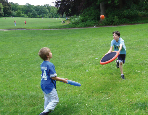 Zwei Jungen spielen mit Fangscheiben und einem Ball auf einer Wiese. Im Hintergrund sieht man weitere Menschen im Park.