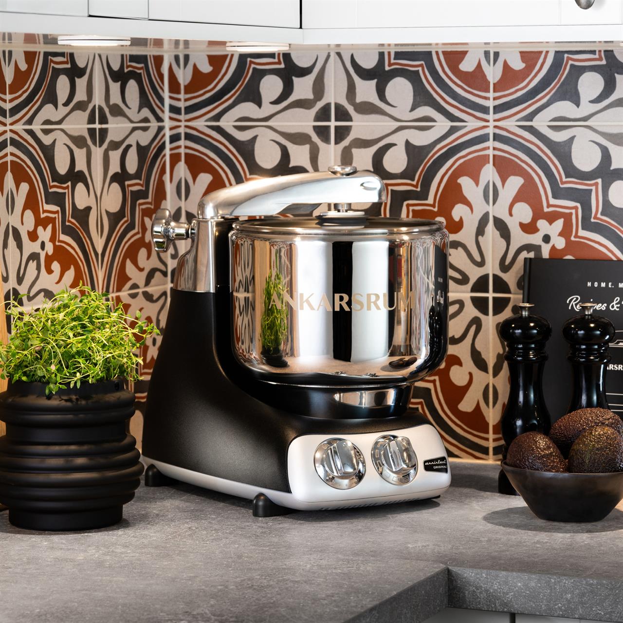 Die Küchenmaschine Black steht auf einer Küchenarbeitsplatte vor einer gemusterten Wandfliese. Neben ihr stehen ein Kräutertopf, ein Salz- und Pfefferstreuer und eine Schale mit Avocados.