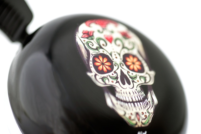 Die Abbildung zeigt eine schwarze Fahrradklingel mit einem bunten Totenkopf im Sugar Skull Stil. Die Verzierung ist farbenfroh und detailreich gestaltet.