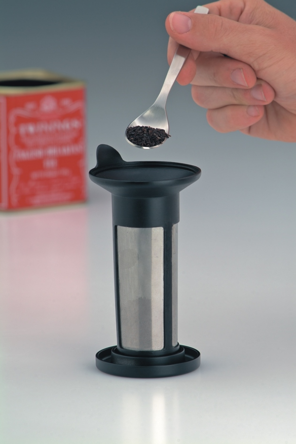 Eine Hand hält einen Löffel mit Teeblättern über einen Teefilter. Im Hintergrund steht eine rote Teedose.