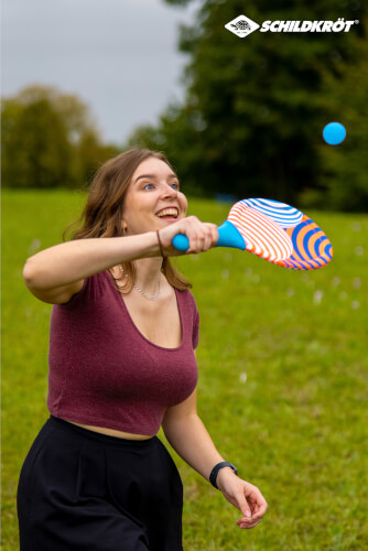 Eine junge Frau spielt mit einem bunten Schläger und einem blauen Ball im Freien. Sie sieht glücklich aus und befindet sich auf einer Wiese.