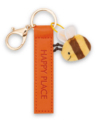 Auf dem Bild ist ein Schlüsselanhänger mit einem orangefarbenen Anhänger, auf dem "HAPPY PLACE" steht. Daran hängt ein kleiner, plüschiger Bienenanhänger.