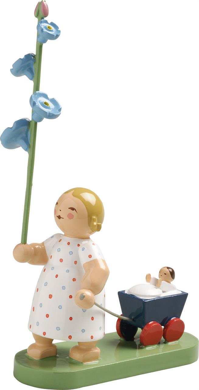 Das Bild zeigt eine Holzfigur eines Mädchens im weißen Kleid mit bunten Punkten, das eine große blaue Blume hält. Neben ihr zieht sie einen kleinen Wagen mit einem Baby darin.