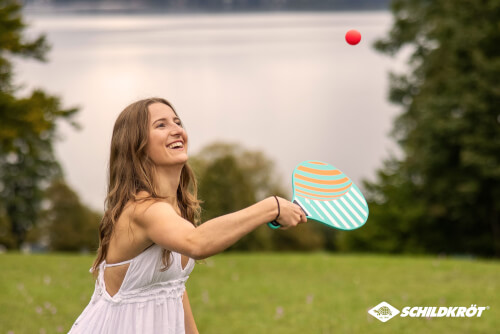 Eine junge Frau spielt mit einem bunten Schläger und einem roten Ball draußen auf einer Wiese. Im Hintergrund ist ein See und grüne Bäume zu sehen.