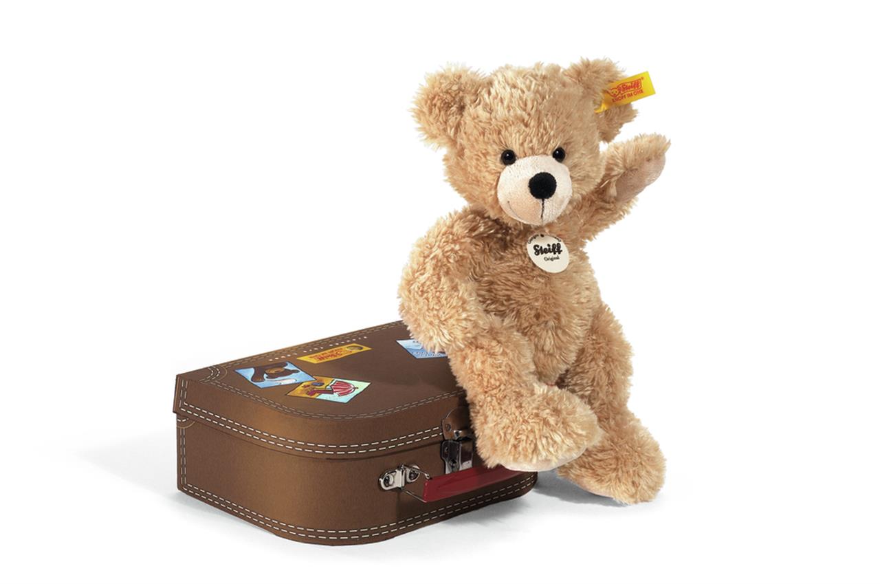 Das Bild zeigt einen braunen Teddybär, der "Fynn" heißt und 28 cm groß ist. Der Teddybär sitzt auf einem kleinen braunen Koffer mit bunten Aufklebern.