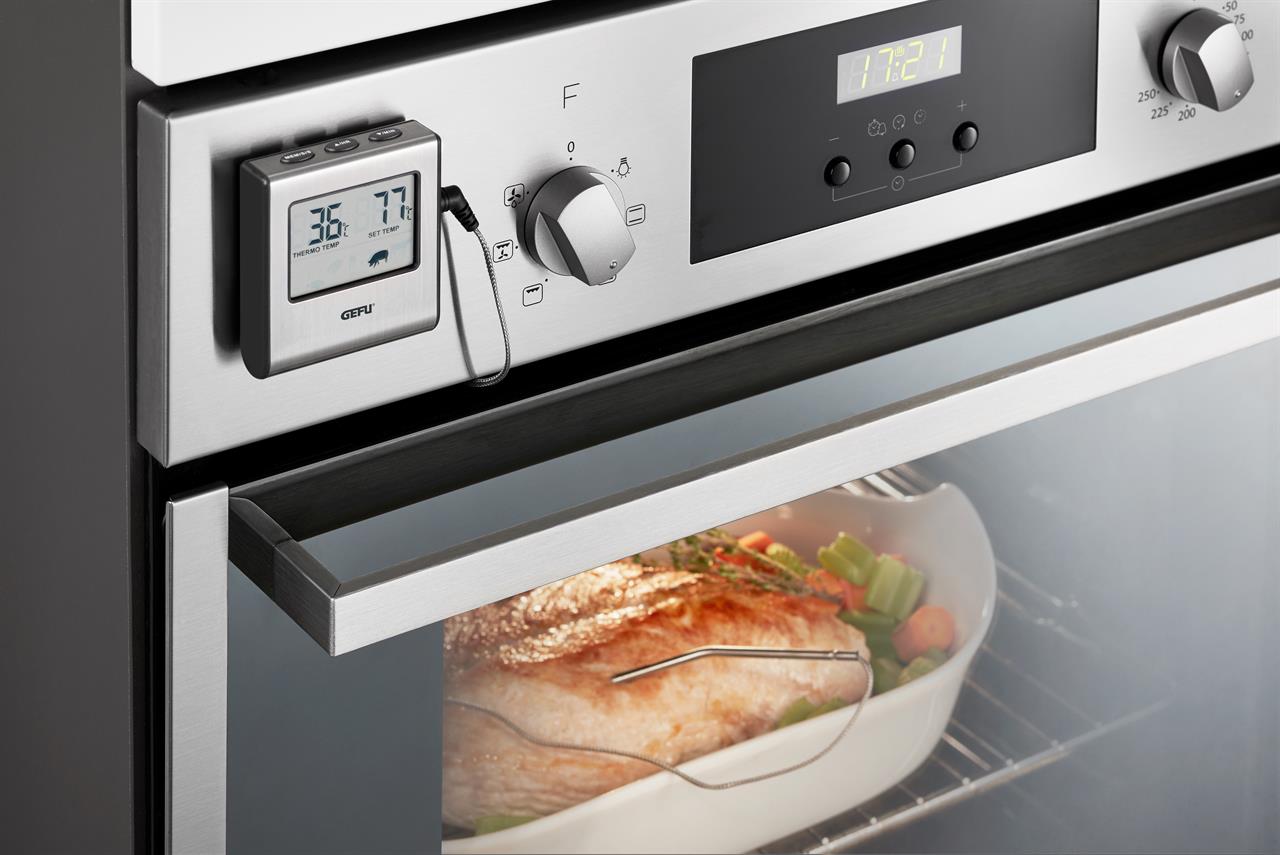 Das Bild zeigt ein digitales Bratenthermometer, das an einem Ofen angebracht ist. Im Ofen wird ein Braten mit Gemüsestücken in einer Auflaufform zubereitet, während das Thermometer die Temperatur misst.