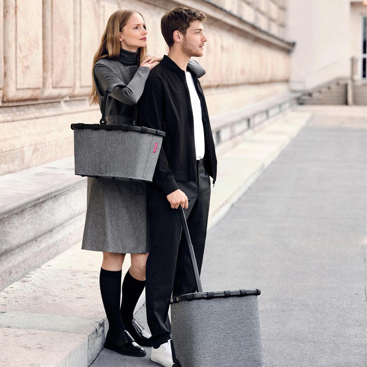 Eine Frau und ein Mann stehen zusammen auf einem Gehweg, beide halten einen "carrycruiser frame twist silver" in der Hand. Die Körbe haben ein modernes, graues Design und wirken praktisch für unterwegs.