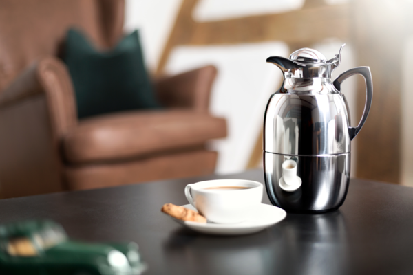 Auf einem Tisch stehen eine Tasse Kaffee mit einem Keks und eine glänzende Edelstahlkanne. Im Hintergrund ist unscharf ein brauner Sessel zu sehen.