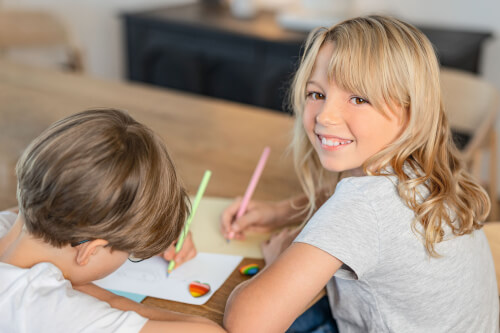 Zwei Kinder sitzen am Tisch und malen mit Buntstiften. Das Mädchen lächelt fröhlich in die Kamera.