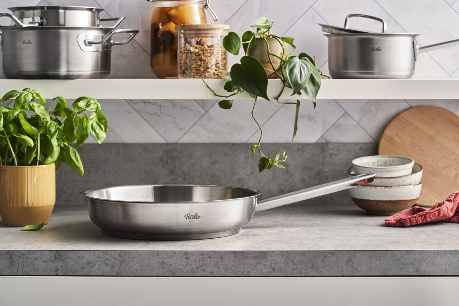 Auf einer Küchenarbeitsplatte steht eine Edelstahlpfanne von Fissler. Im Hintergrund befinden sich Kräuter, Geschirr, Töpfe und einige Pflanzen.