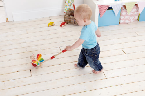 Ein kleines Kind zieht ein buntes Holzspielzeug auf Rädern hinter sich her. Es läuft barfuß über einen hellen Holzboden.