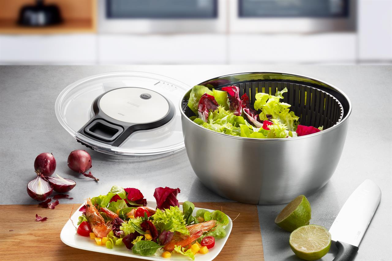 Die Salatschleuder SPEEDWING®, Edelstahl, steht auf einer Küchenarbeitsplatte, gefüllt mit frischem Salat. Daneben liegen ihr Deckel, ein Messer, Zwiebeln und ein Teller mit angerichteter Salatmischung.