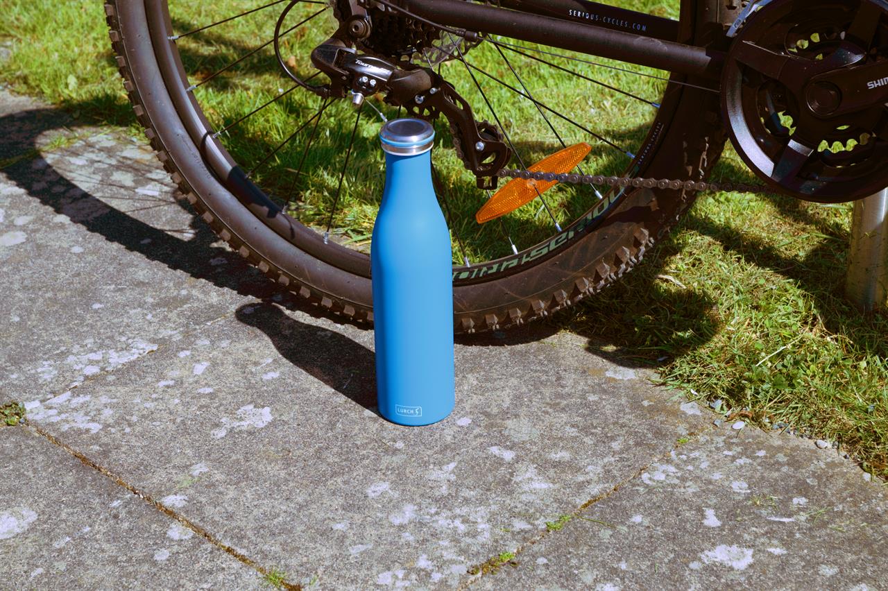 Die Isolier-Flasche Edelstahl 0,75l Azur steht auf einem Gehweg neben einem Fahrrad. Sie ist leuchtend blau und hat einen silbernen Deckel.