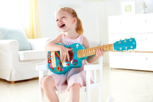Ein kleines Mädchen sitzt lachend auf einem weißen Stuhl und spielt eine bunte, blaue Gitarre. Sie wirkt glücklich und genießt den Moment im hellen Wohnzimmer.