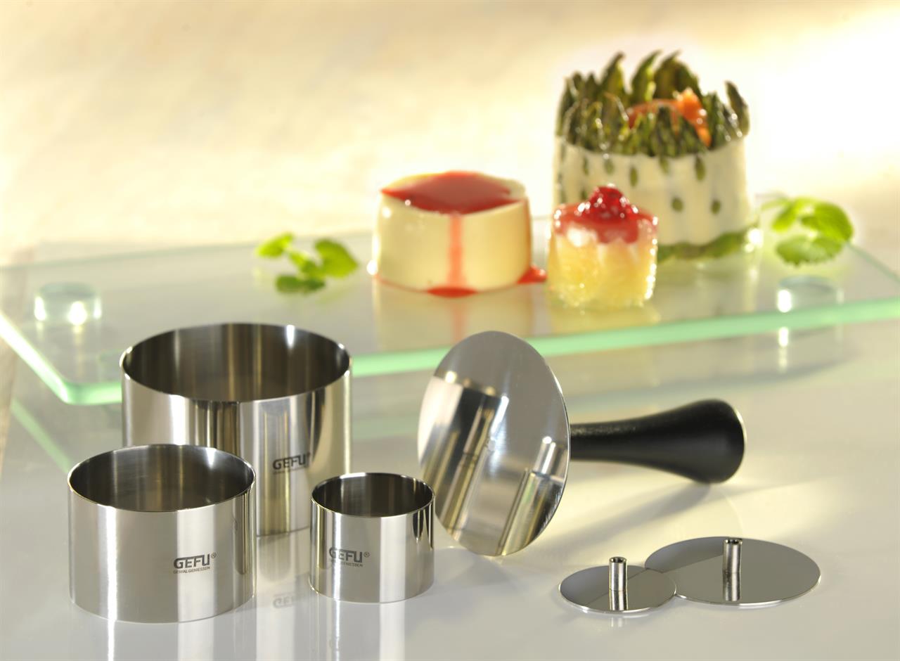 Das Bild zeigt das Produkt "Profispeisenringe Formidable, 10-tlg." bestehend aus mehreren Metallringen und Zubehör. Im Hintergrund sind attraktiv angerichtete Speisen zu sehen, die mit diesen Ringen geformt wurden.