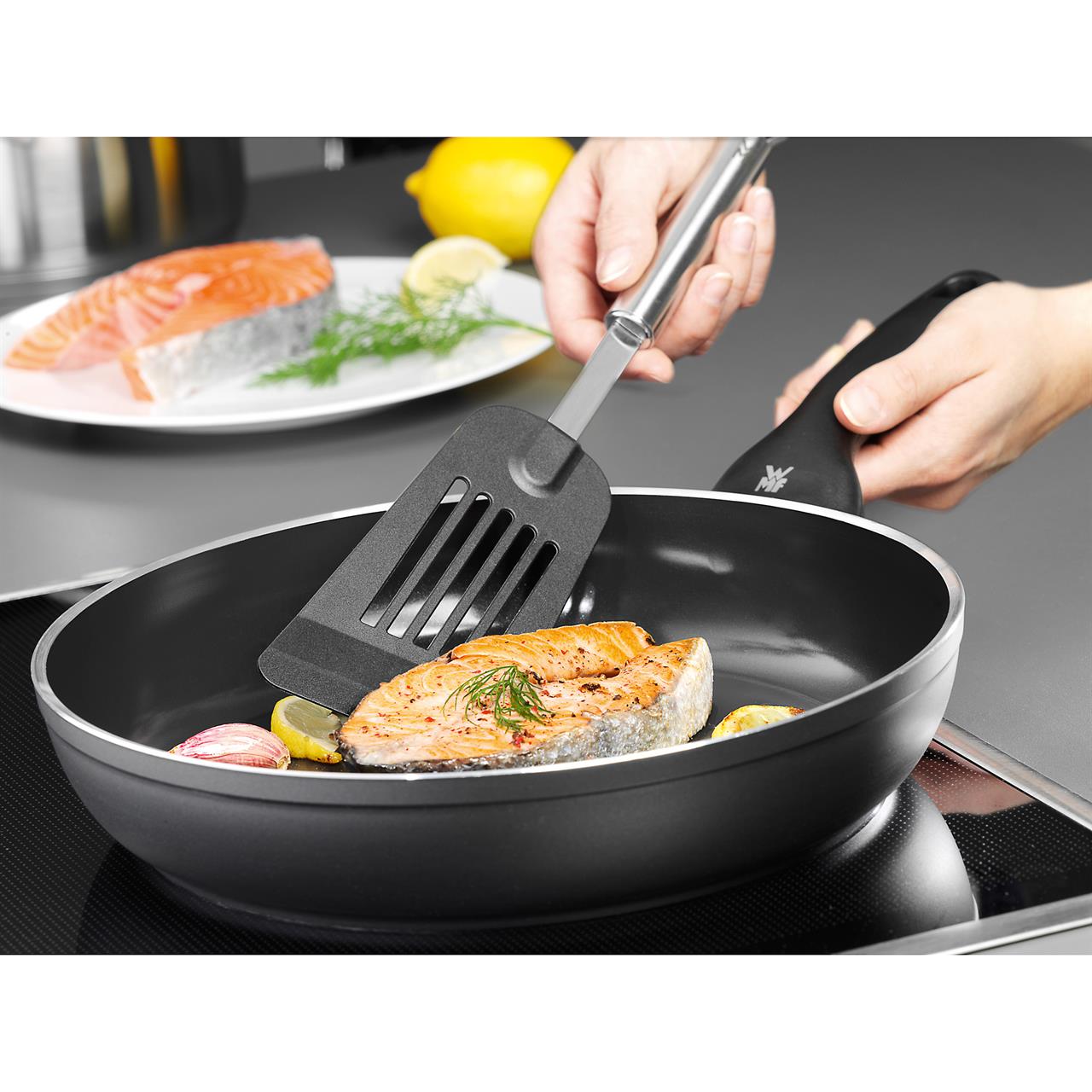 Die Backschaufel Profi Plus wird verwendet, um ein Stück Lachs in einer Pfanne zu wenden. Im Hintergrund sieht man ein weiteres Stück Lachs auf einem Teller mit Zitrone und Dill.