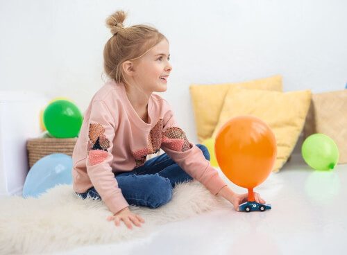 Ein Mädchen sitzt auf einem Teppich und spielt mit einem Auto, das einen orangefarbenen Ballon hat. Im Hintergrund liegen bunte Luftballons und Kissen auf dem Boden.