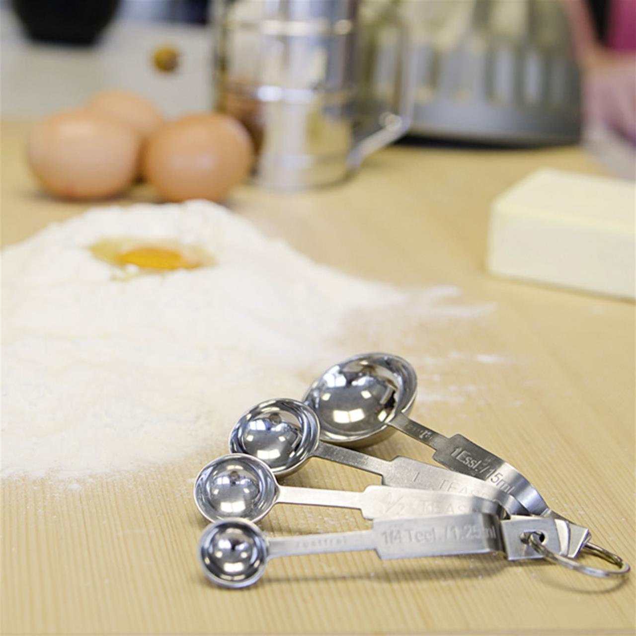 Im Vordergrund liegt ein Messlöffel-Set aus Metall auf einer Arbeitsfläche. Im Hintergrund sind Mehl, ein Ei, Eier und ein Stück Butter zu sehen, was auf eine Backvorbereitung hindeutet.