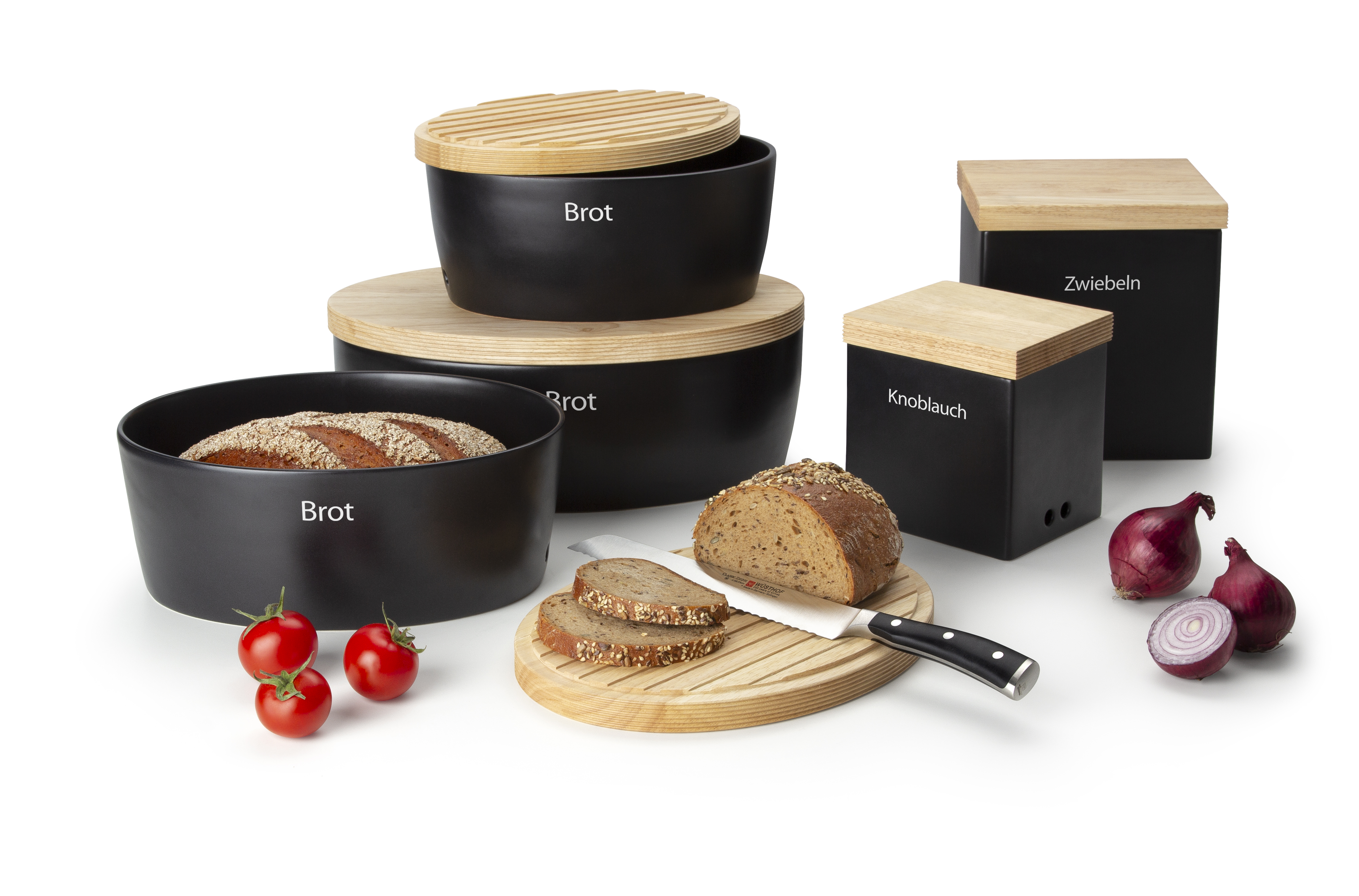 Auf dem Bild sind verschiedene schwarze Vorratsdosen mit Holzdeckeln zu sehen, die mit „Brot“, „Knoblauch“ und „Zwiebeln“ beschriftet sind. Vor den Dosen liegen ein aufgeschnittenes Brot, ein Brotmesser, Tomaten und rote Zwiebeln.