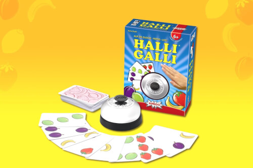 Auf dem Bild ist das Kartenspiel "Halli Galli" mit Spielkarten, einer Glocke und der Spielverpackung zu sehen. Die Karten zeigen bunte Früchte und sind vor einer gelben Hintergrundfläche ausgebreitet.