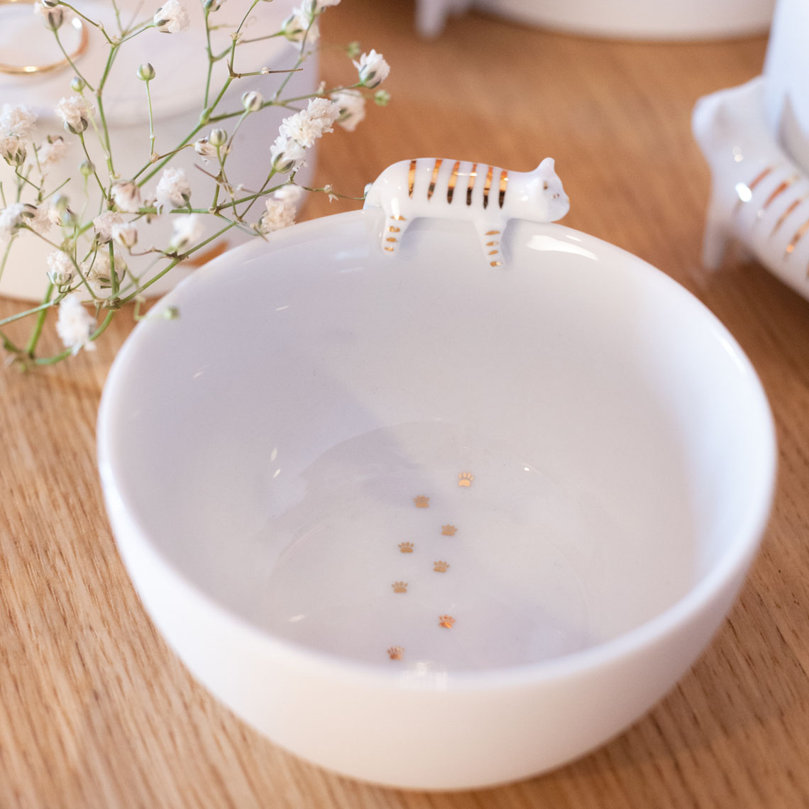 Eine weiße Schale steht auf einem Holztisch. Am Rand sitzt eine kleine Keramikfigur einer Katze und auf dem Schalenboden sind goldene Pfotenabdrücke.