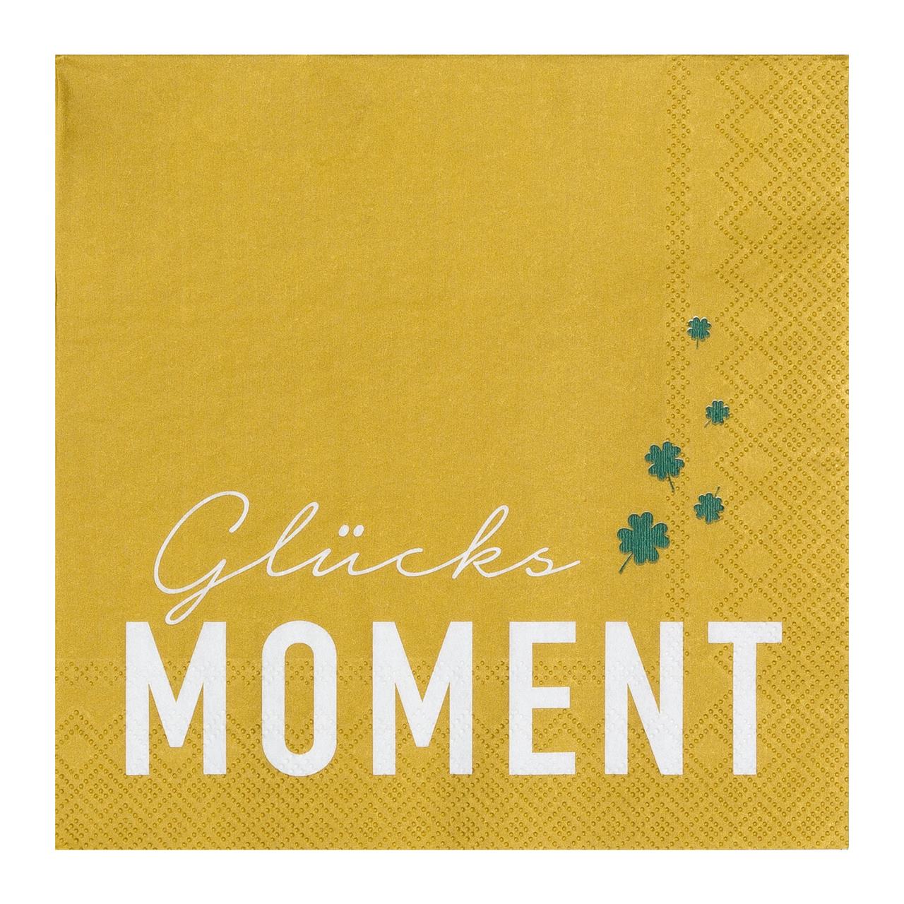 Die Serviette zeigt die Aufschrift „Glücksmoment“ in weißer Schrift auf gelbem Hintergrund. Rechts sind einige kleine grüne Kleeblätter abgebildet.
