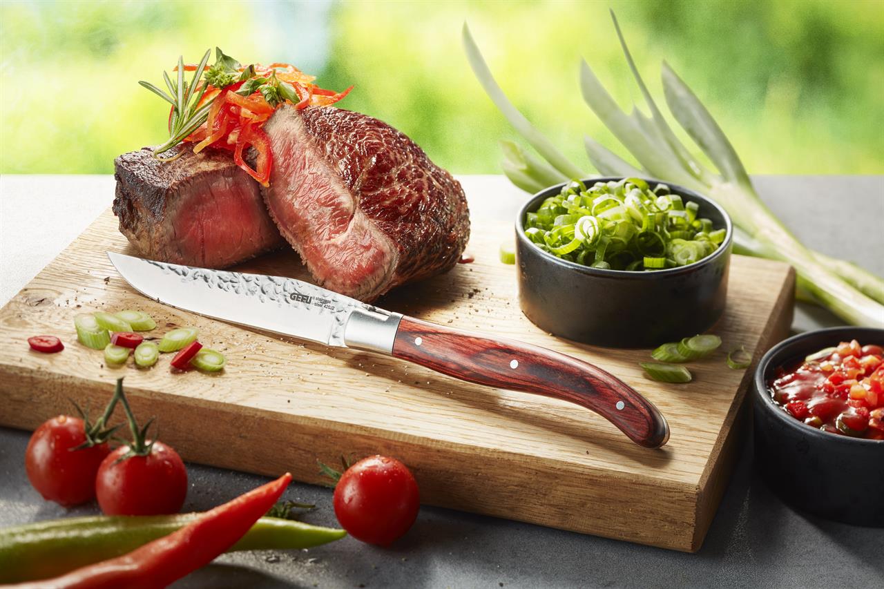Auf einem Holzbrett liegt ein Stück saftiges Steak, das mit einem Steakmesser aus dem Set "Steakmesser-Set RANCHO, 6 Stück" angeschnitten wurde. Rundherum sind frische Zutaten wie Tomaten, Frühlingszwiebeln und ein Schälchen Salsa zu sehen.