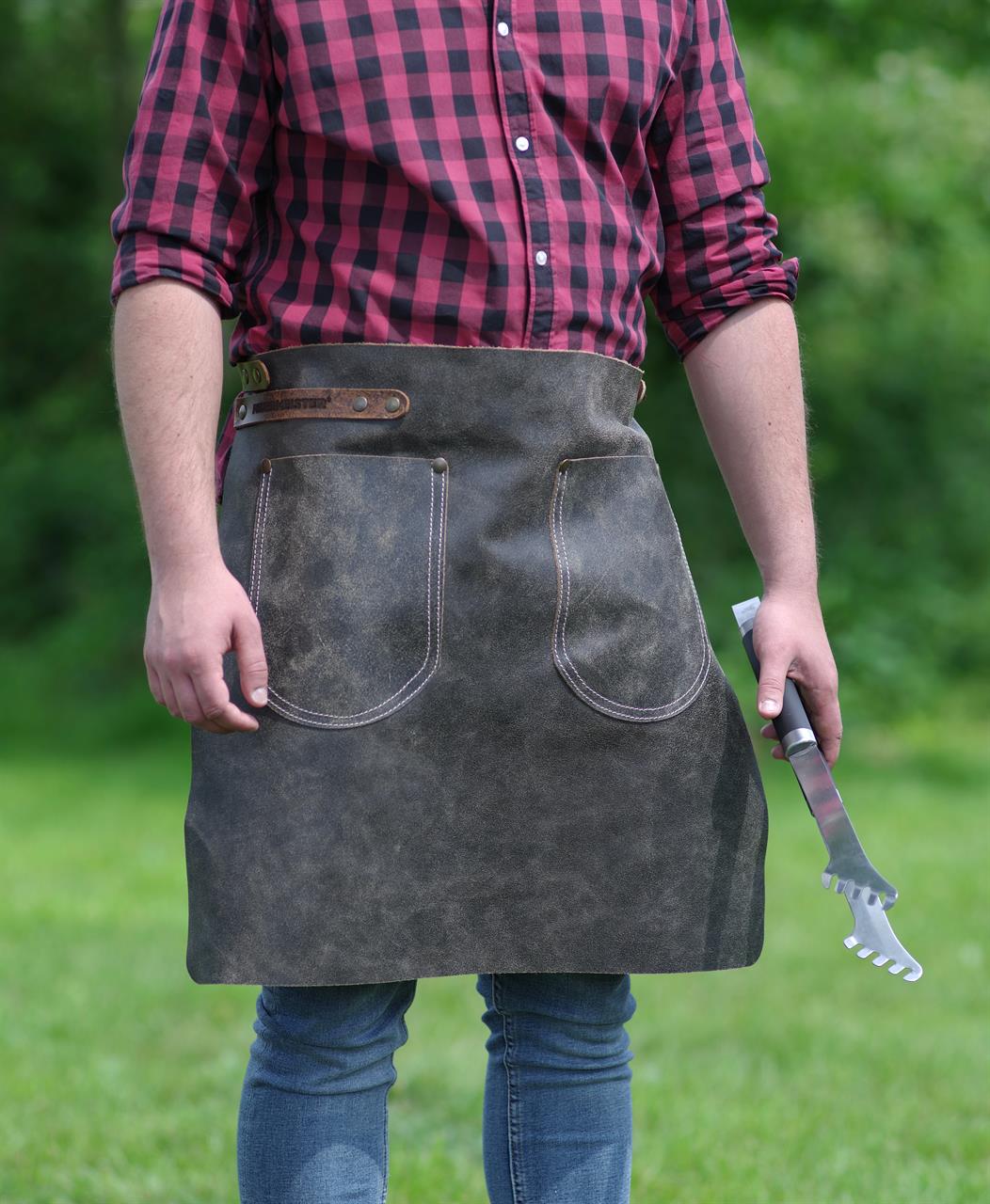 Eine Person trägt eine Hüftschürze aus antik aussehendem Leder in der Größe 2 (70x55 cm) in der Farbe Anthrazit. Die Schürze hat zwei große Taschen auf der Vorderseite und wird im Freien getragen.