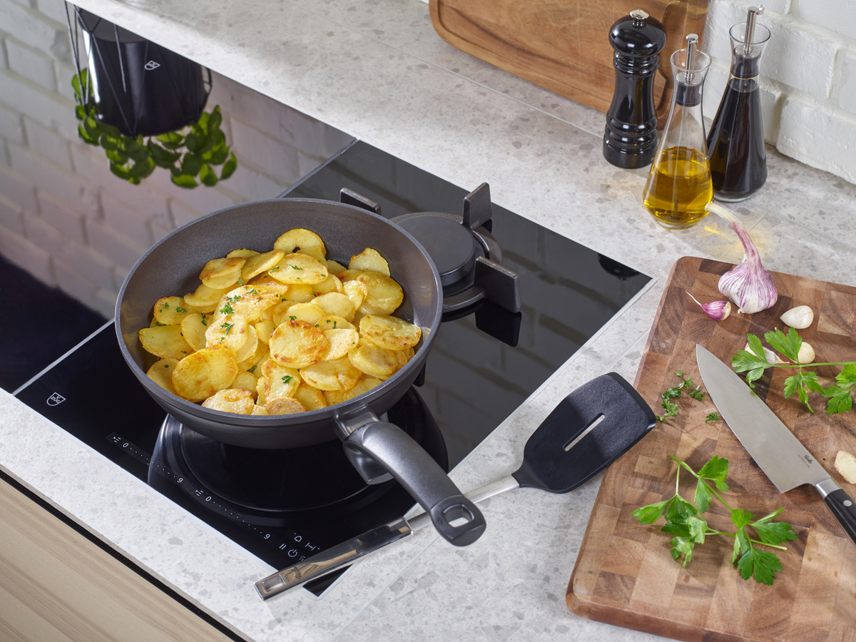 In der Pfanne werden goldbraune, gebratene Kartoffelscheiben zubereitet. Daneben liegen auf dem Schneidebrett frische Kräuter, Knoblauch und ein Messer.