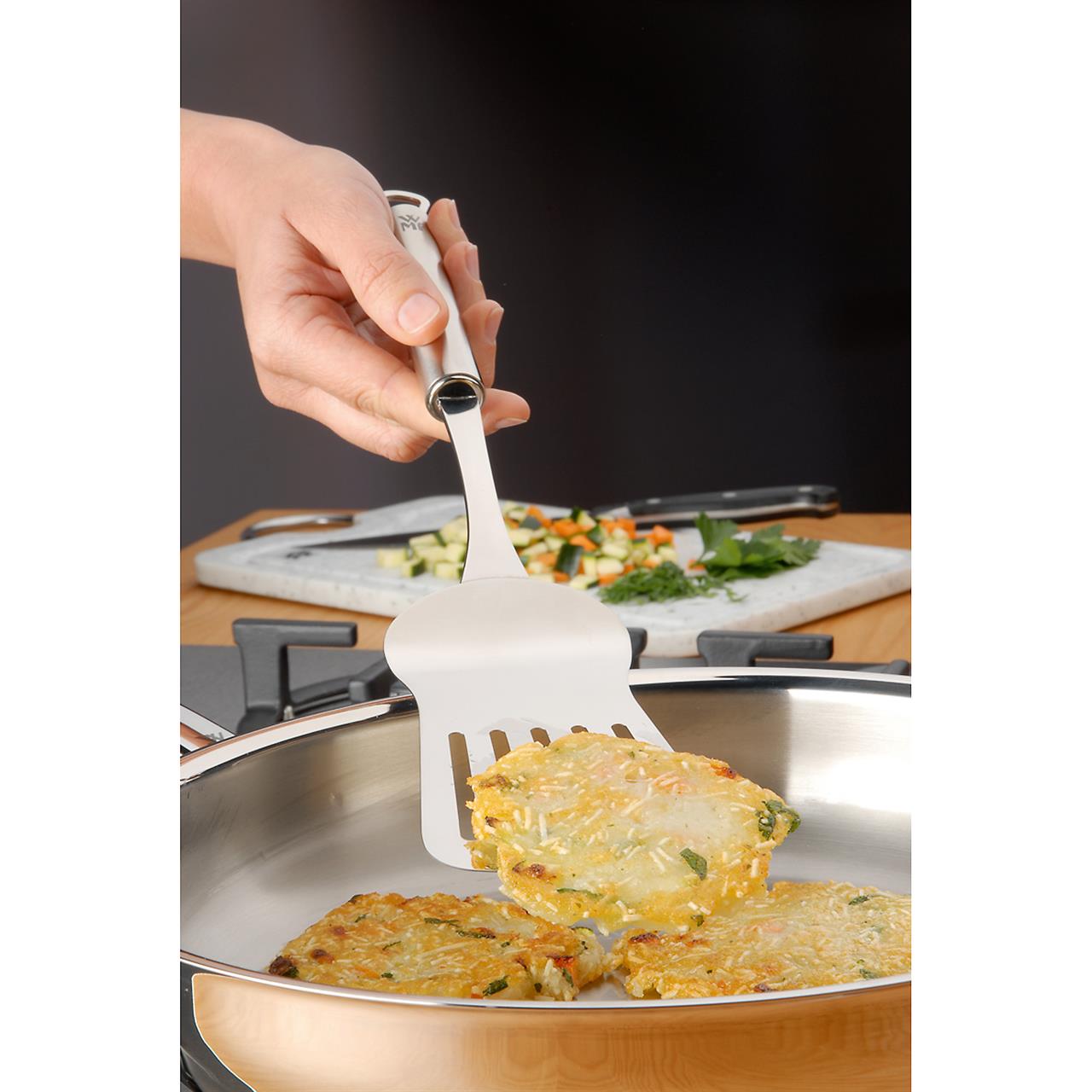 Eine Hand hält die Backschaufel Profi Plus und hebt damit einen Kartoffelpuffer aus einer Pfanne. Im Hintergrund liegt auf einem Schneidebrett geschnittenes Gemüse bereit.