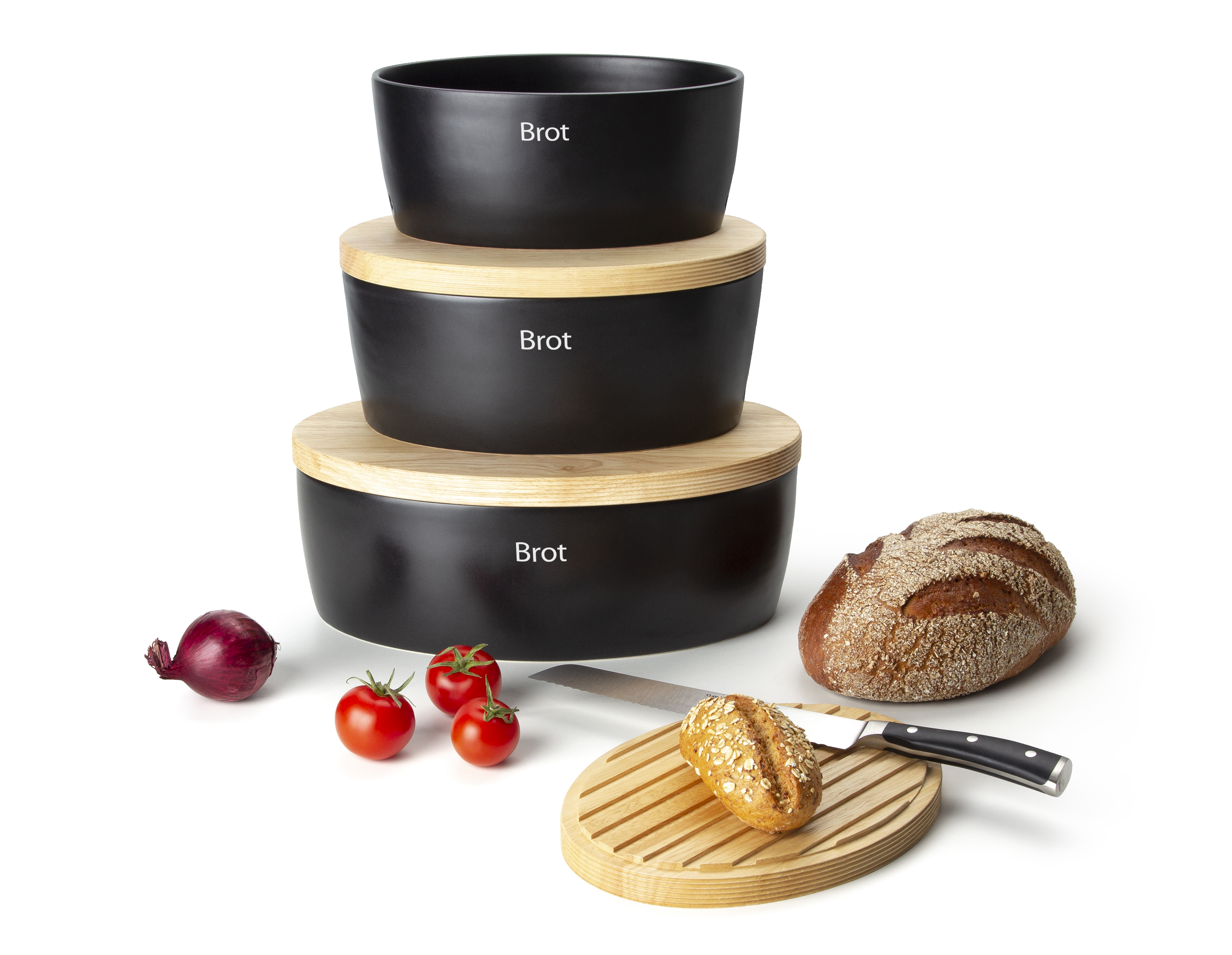 Auf dem Bild sind drei schwarze Brotdosen mit Holzdeckeln zu sehen, auf denen jeweils das Wort "Brot" steht. Daneben liegen ein Laib Brot, ein Brötchen auf einem Holzbrett mit Messer, drei Tomaten und eine rote Zwiebel.