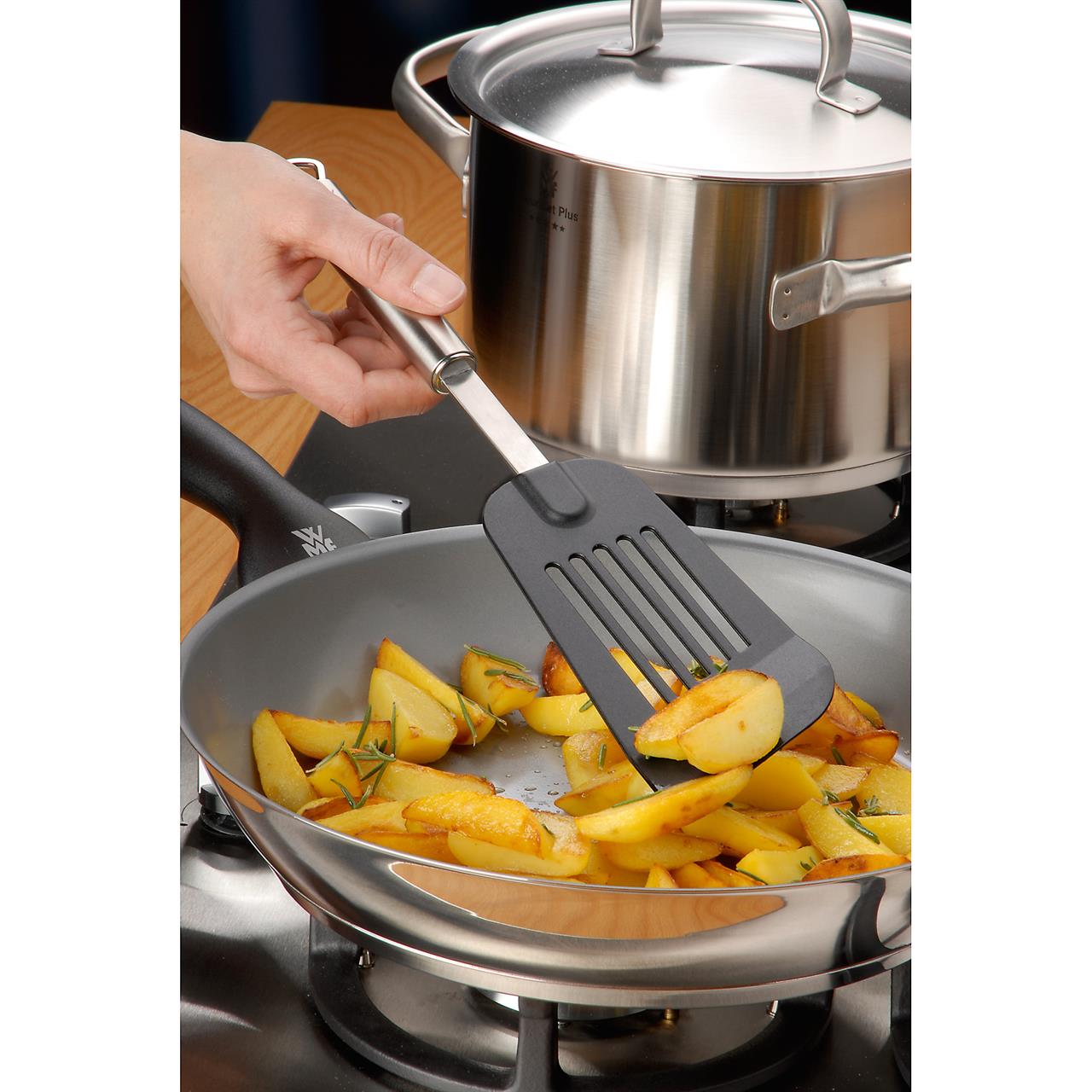 Eine Hand benutzt die Backschaufel Profi Plus, um gebratene Kartoffeln in einer Pfanne zu wenden. Im Hintergrund steht ein Edelstahltopf auf dem Herd.